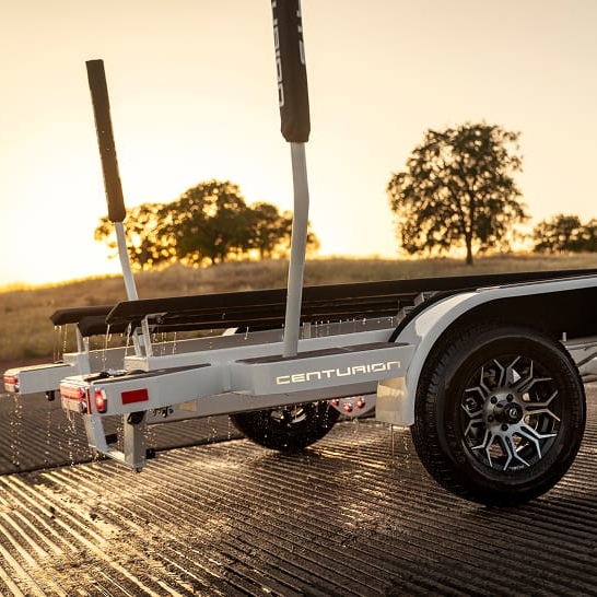 A Centurion Fe Series boat trailer with wet tires is parked on a ramp at sunset, water dripping off and trees visible in the background, highlighting unique personal features against the evening sky.