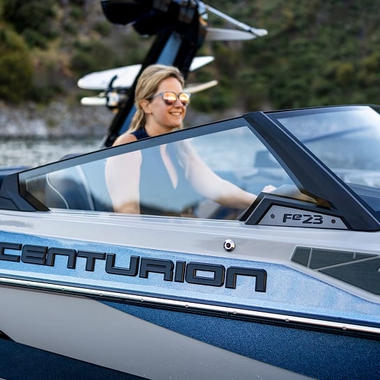 A person with distinct personal features wearing sunglasses smiles while driving a Centurion Fe Series FE23 boat on the water, with trees and hills in the background.