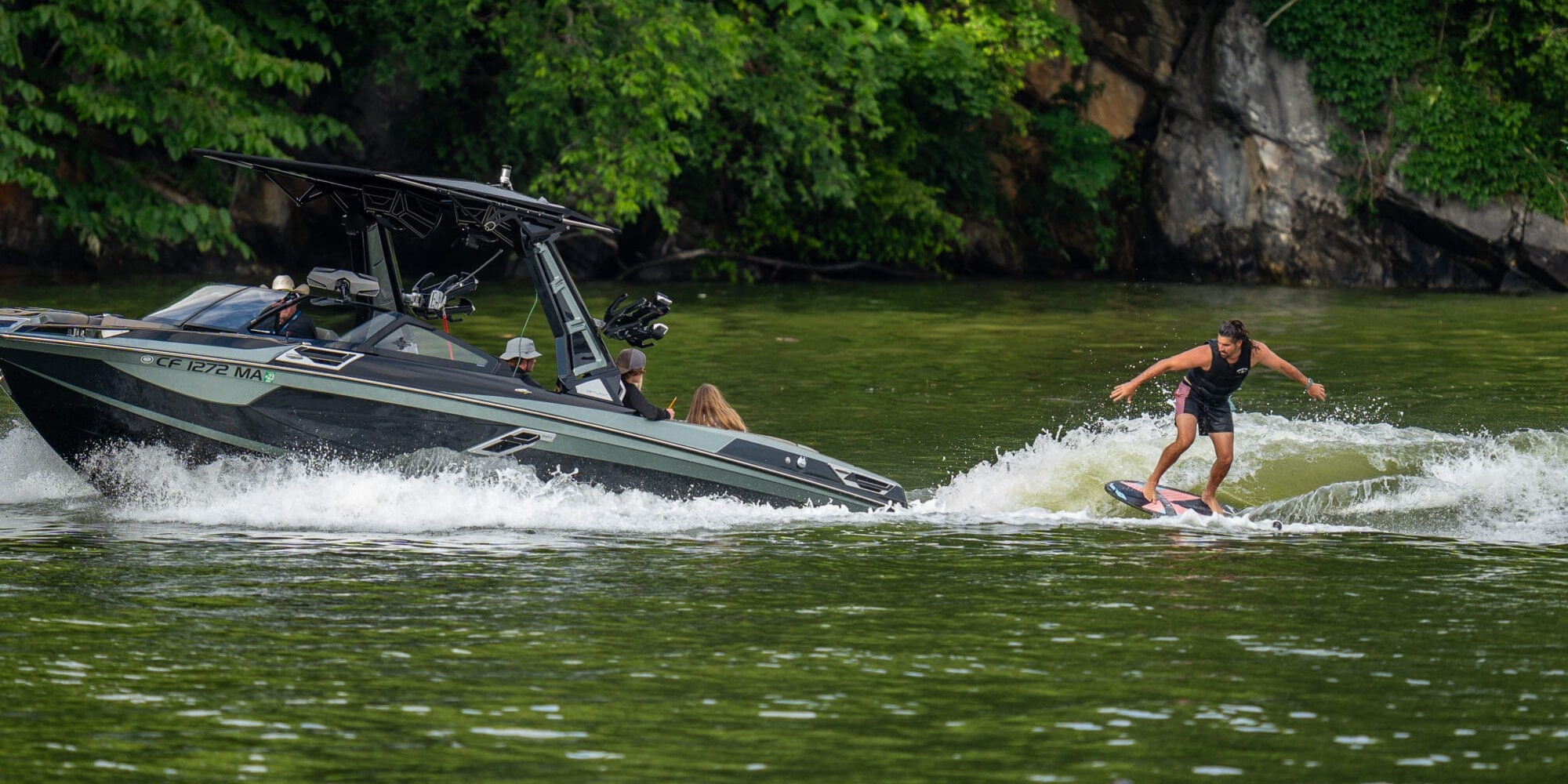 A person is wakesurfing behind a black boat with several passengers on a green, tree-lined body of water.