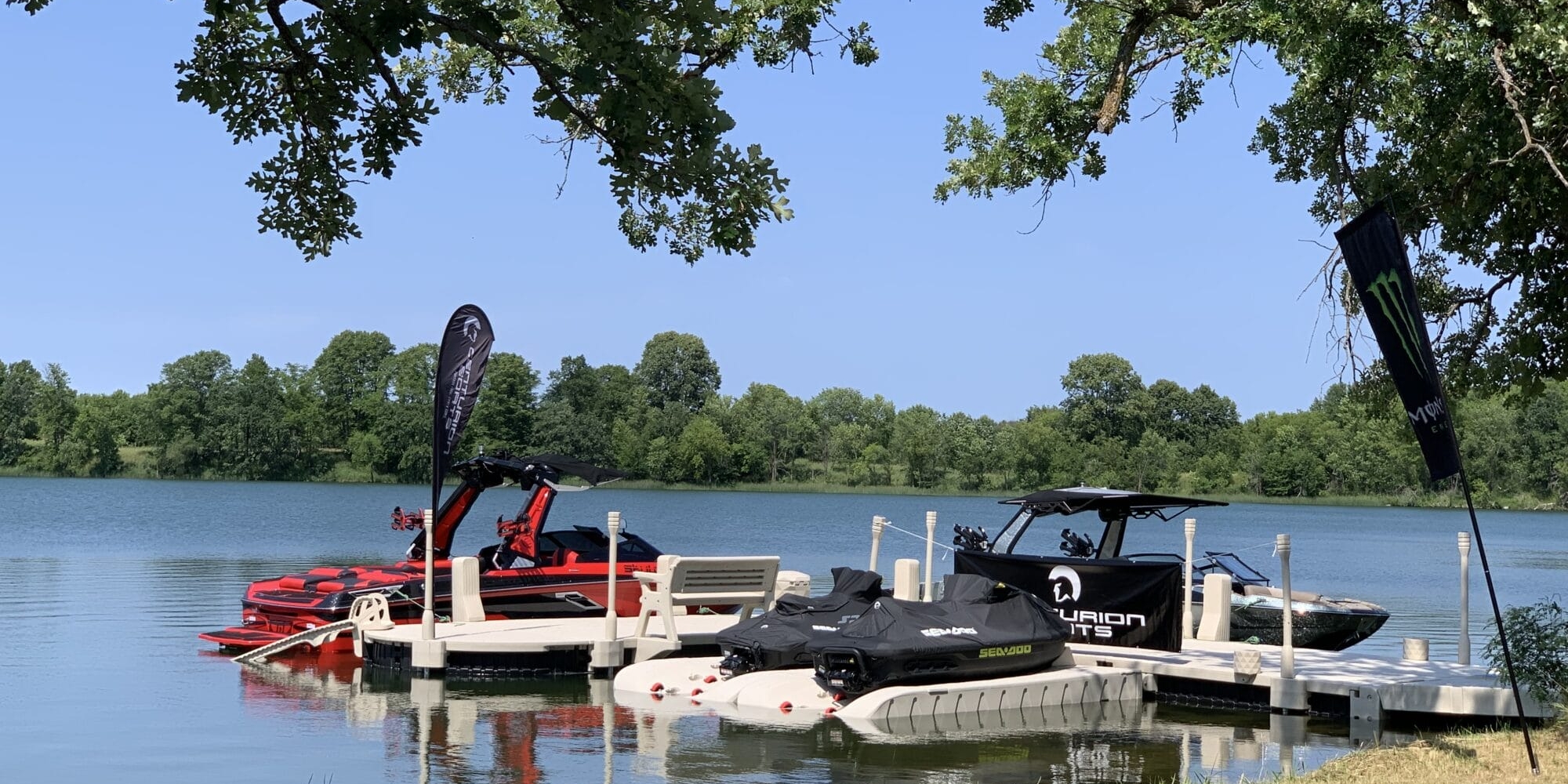 Three boats are docked at a lake shore under a sunny sky. The scene is framed by overhanging tree branches and green grass in the foreground.
