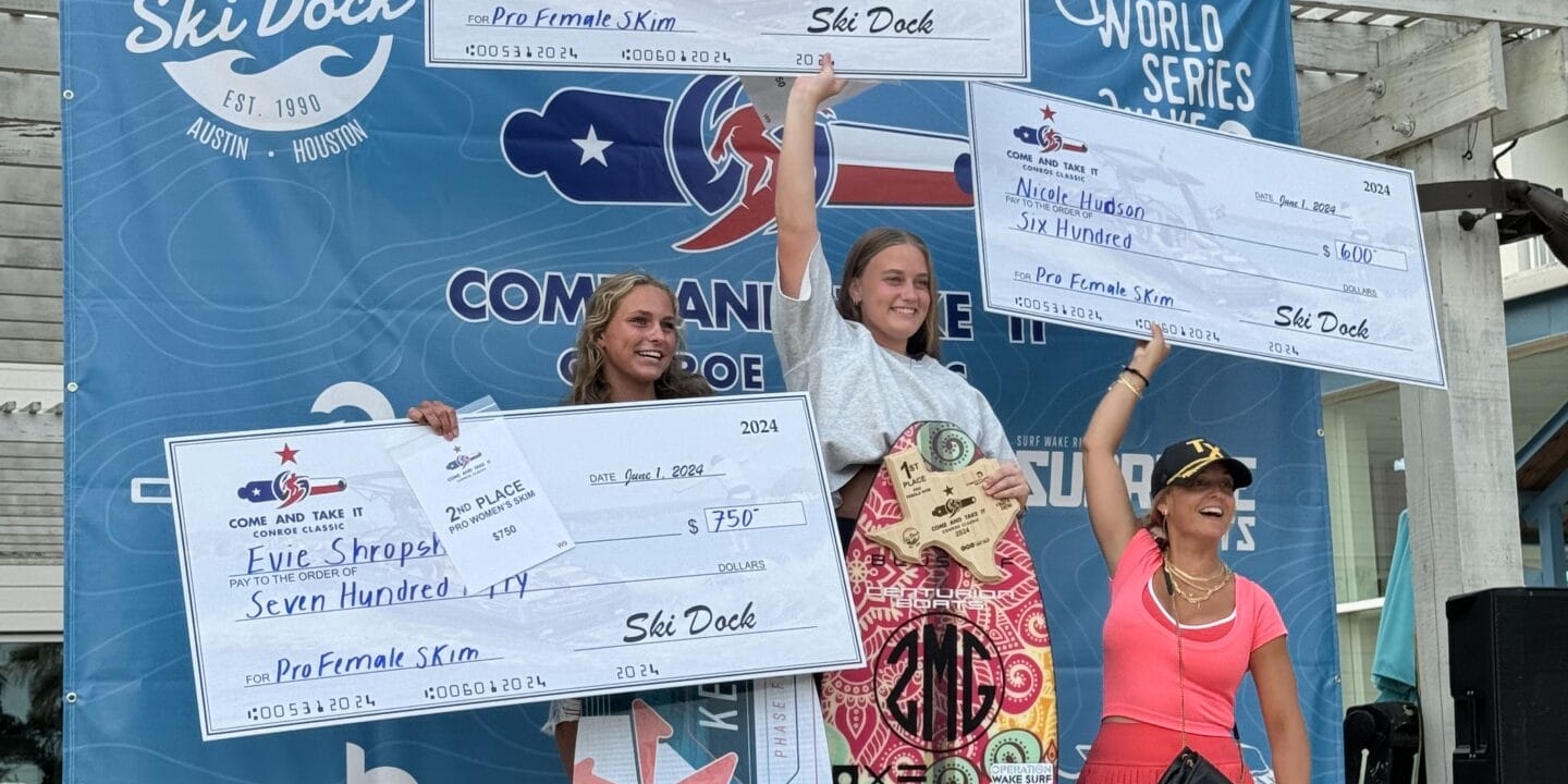 Three female surfers stand on a podium holding large prize checks at a surfing competition. They are smiling, and a surf-themed backdrop displays sponsors' logos.