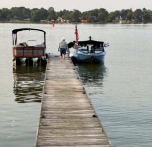 A person walks on a wooden dock toward two moored boats, one pontoon and one small boat with an American flag. A calm lake and houses are visible in the background.