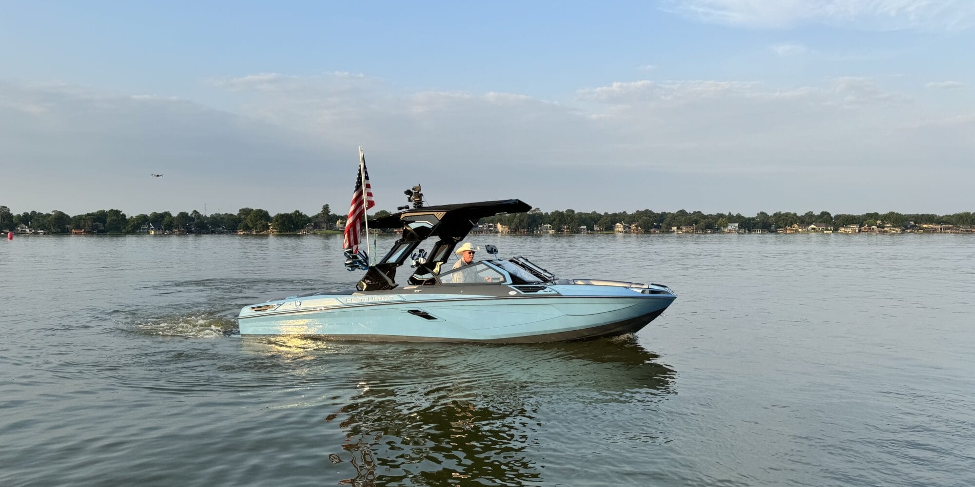 A light blue motorboat with an American flag is moving on a calm lake under a clear sky with some clouds. Trees and houses are visible on the distant shoreline.