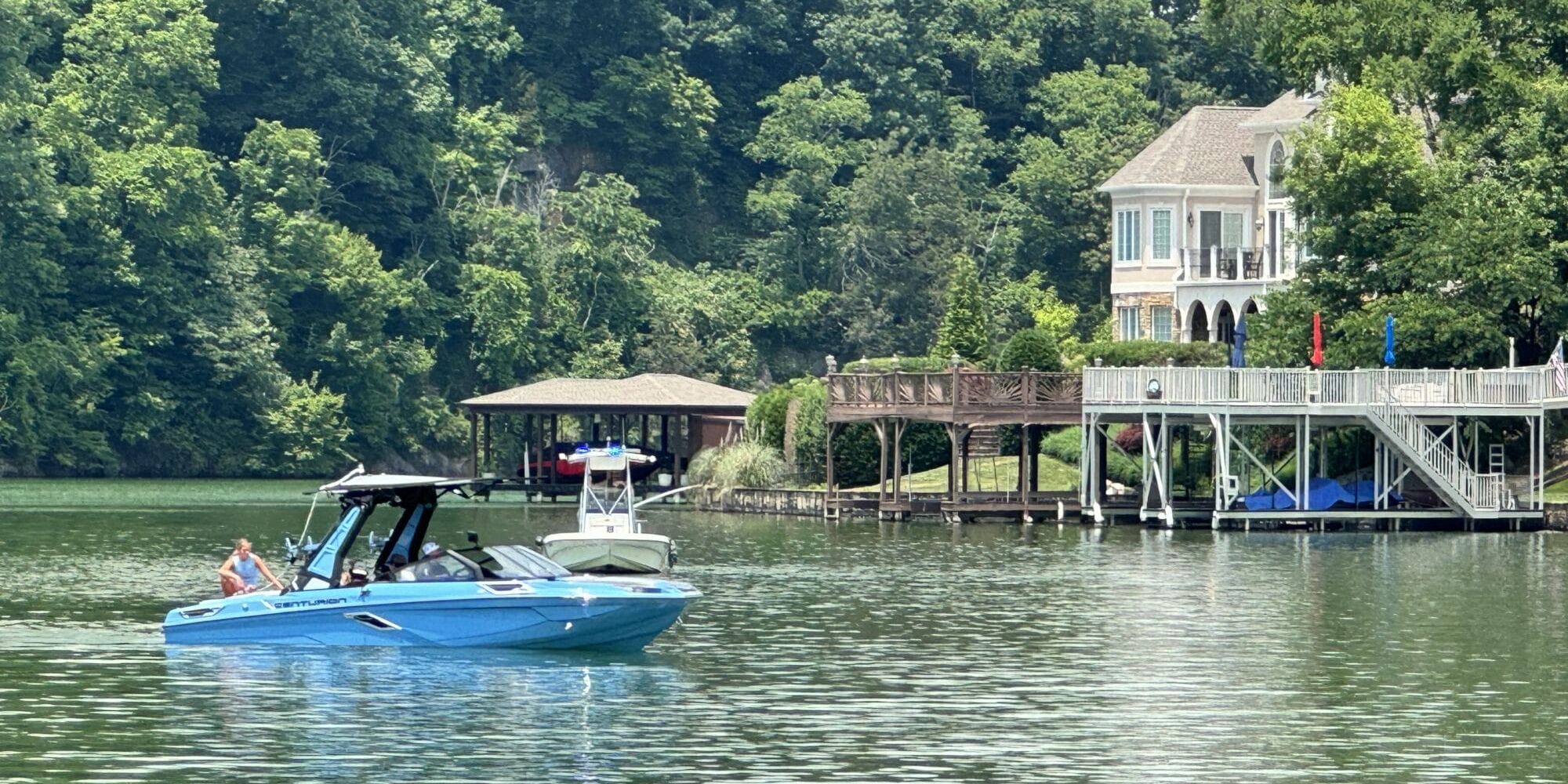 A blue motorboat with several people onboard cruises on a lake near a large lakeside house with a dock and surrounding greenery.