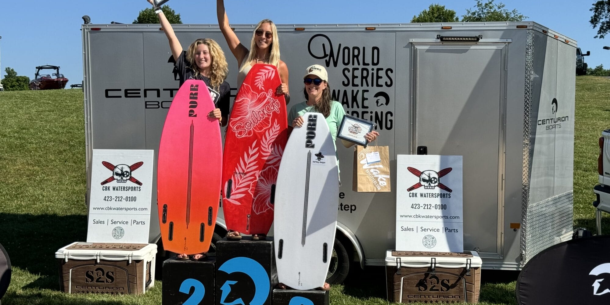 Three individuals stand on a winners podium holding certificates, with two holding surfboards. They are in front of a trailer with 