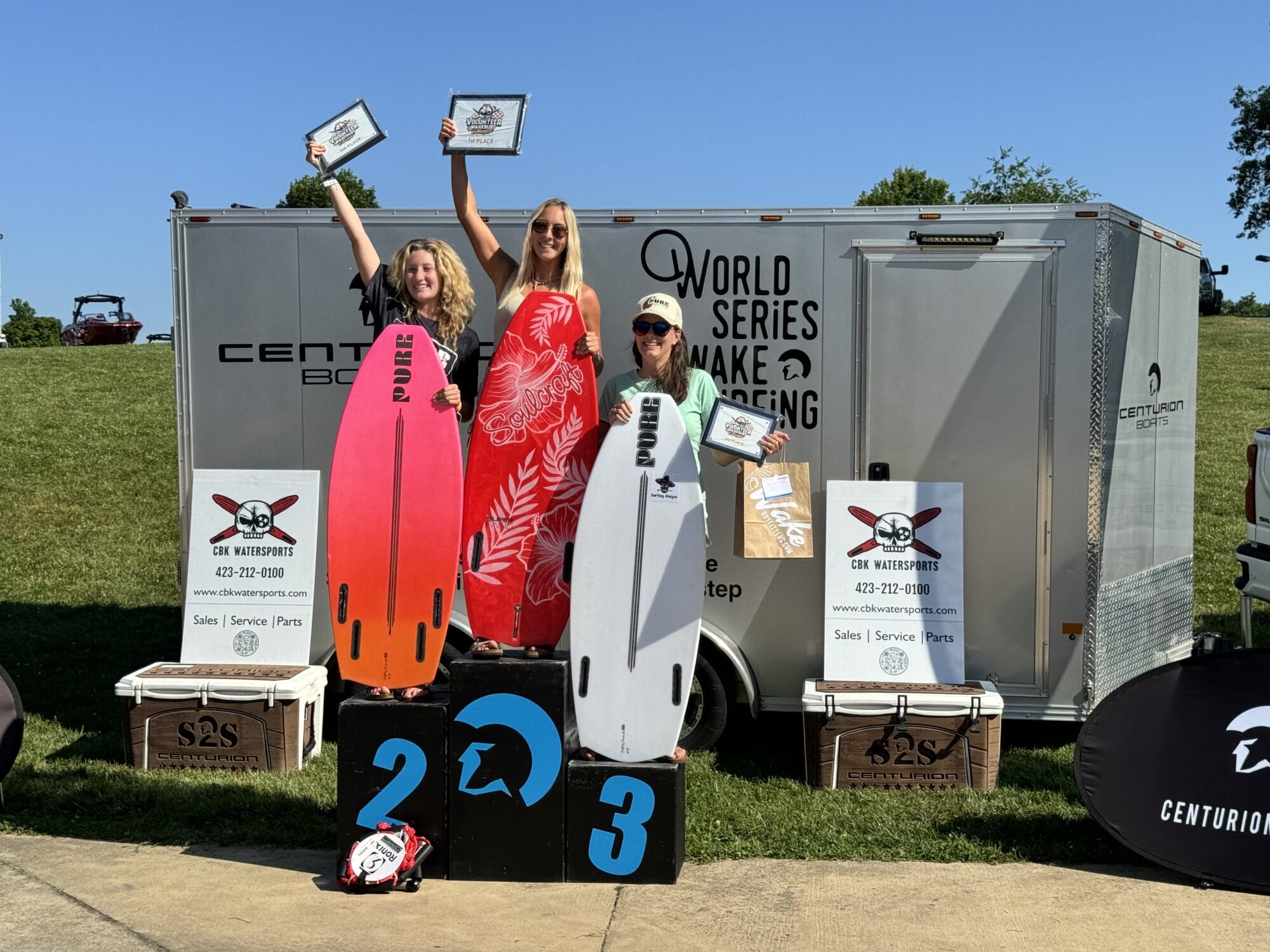 Three individuals stand on a winners podium holding certificates, with two holding surfboards. They are in front of a trailer with "World Series Wake Surfing" signage.