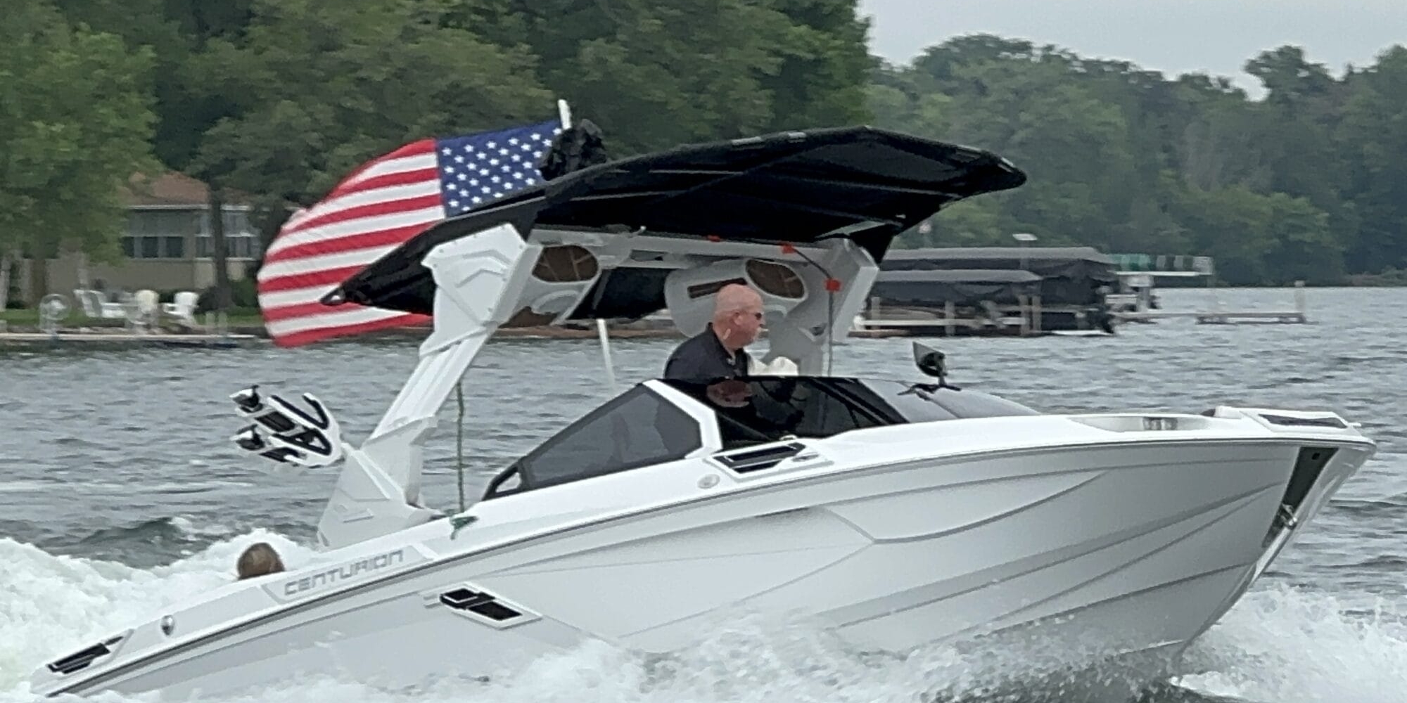 A person is driving a white Centurion boat on a lake. The boat has an American flag at the back, and there are trees and houses visible in the background.
