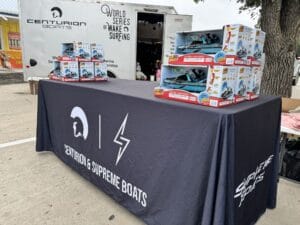 A table covered with a Centurion & Supreme Boats cloth has toys displayed on it. A Centurion Boats trailer and a sign for the World Series of Wake Surfing are in the background.
