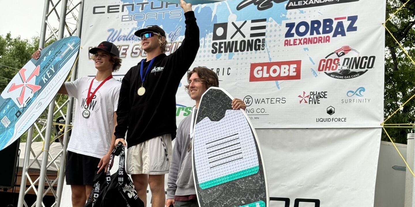 Three individuals stand on a podium holding awards at a surfing competition. The backdrop features various sponsor logos and a banner that reads 