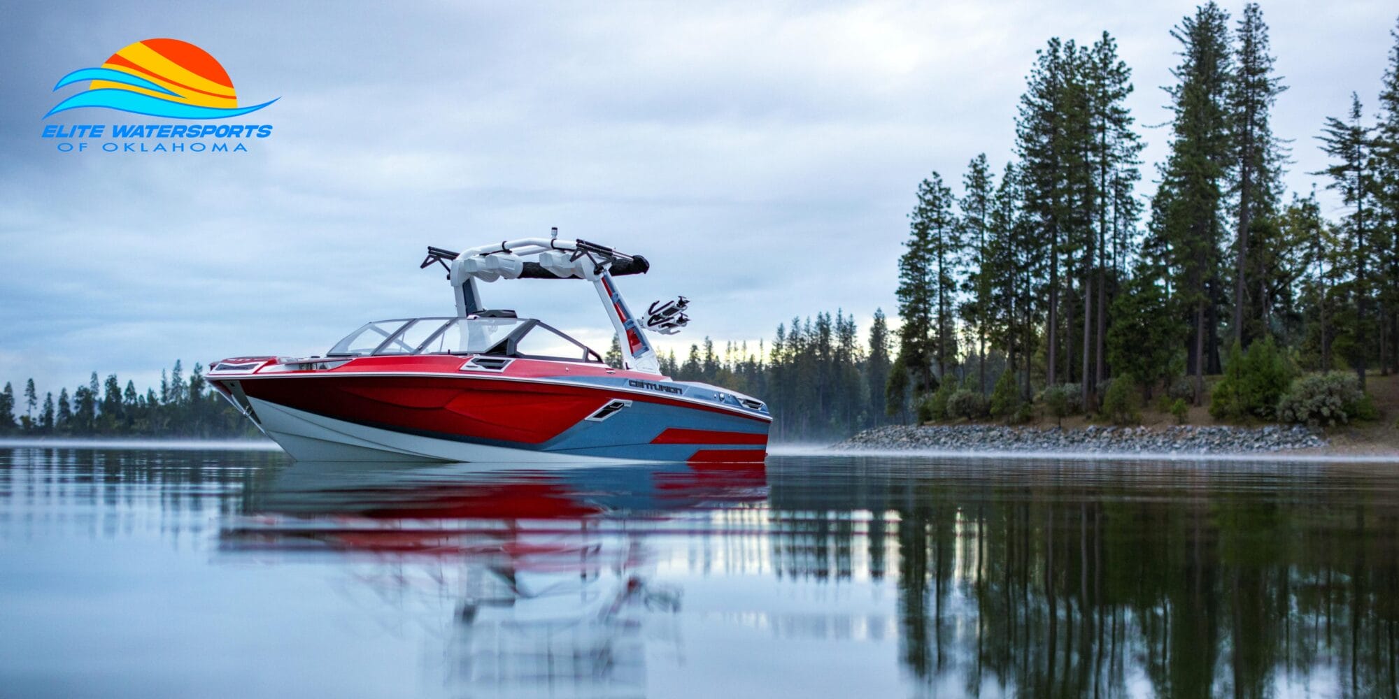 A red and white wakeboard boat is on a calm lake surrounded by trees, with a logo of 
