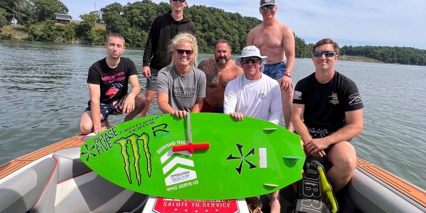 A group of seven people on a boat, one holding a green surfboard. They are on a body of water with trees in the background, under a partly cloudy sky.