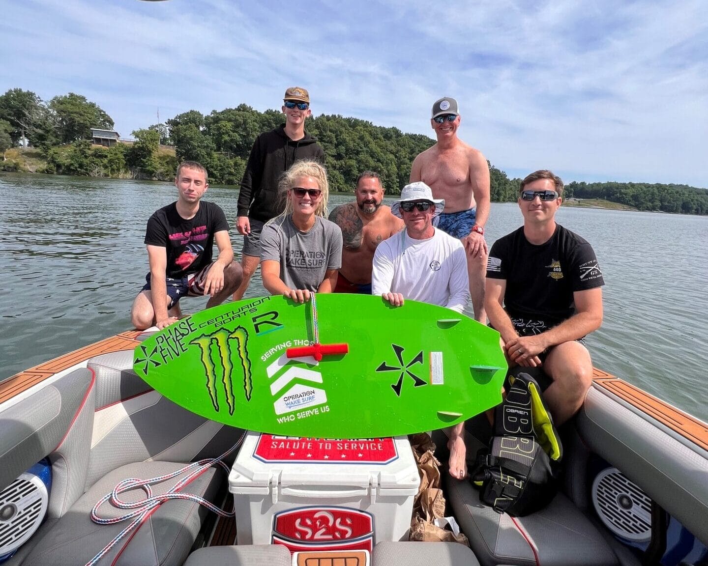A group of seven people on a boat, one holding a green surfboard. They are on a body of water with trees in the background, under a partly cloudy sky.