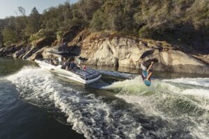 A person wakeboarding performs a flip while being towed by a motorboat near a rocky shoreline.