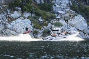 A person rides a wakeboard on the water, propelled by a motorboat with several people on board, near a rocky shoreline.