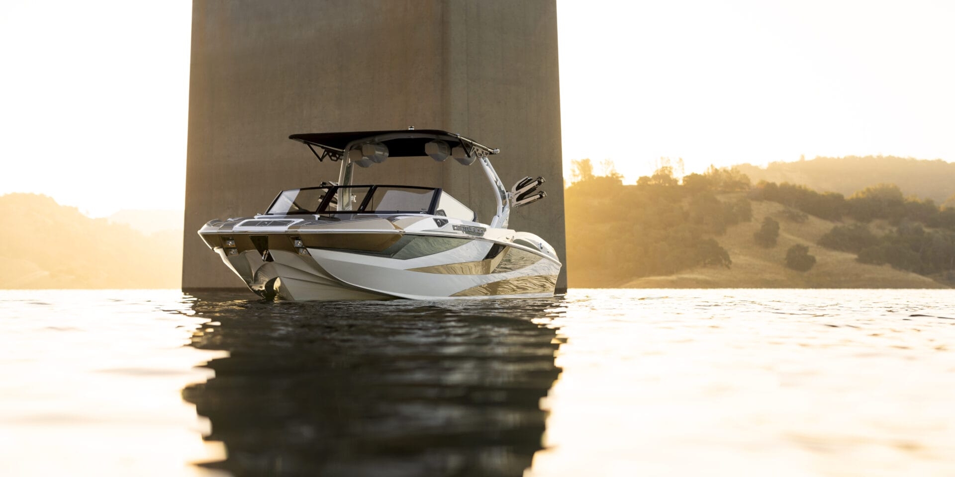 A motorboat anchored underneath a tall bridge pillar, with calm water reflecting the sunset and distant hills in the background.