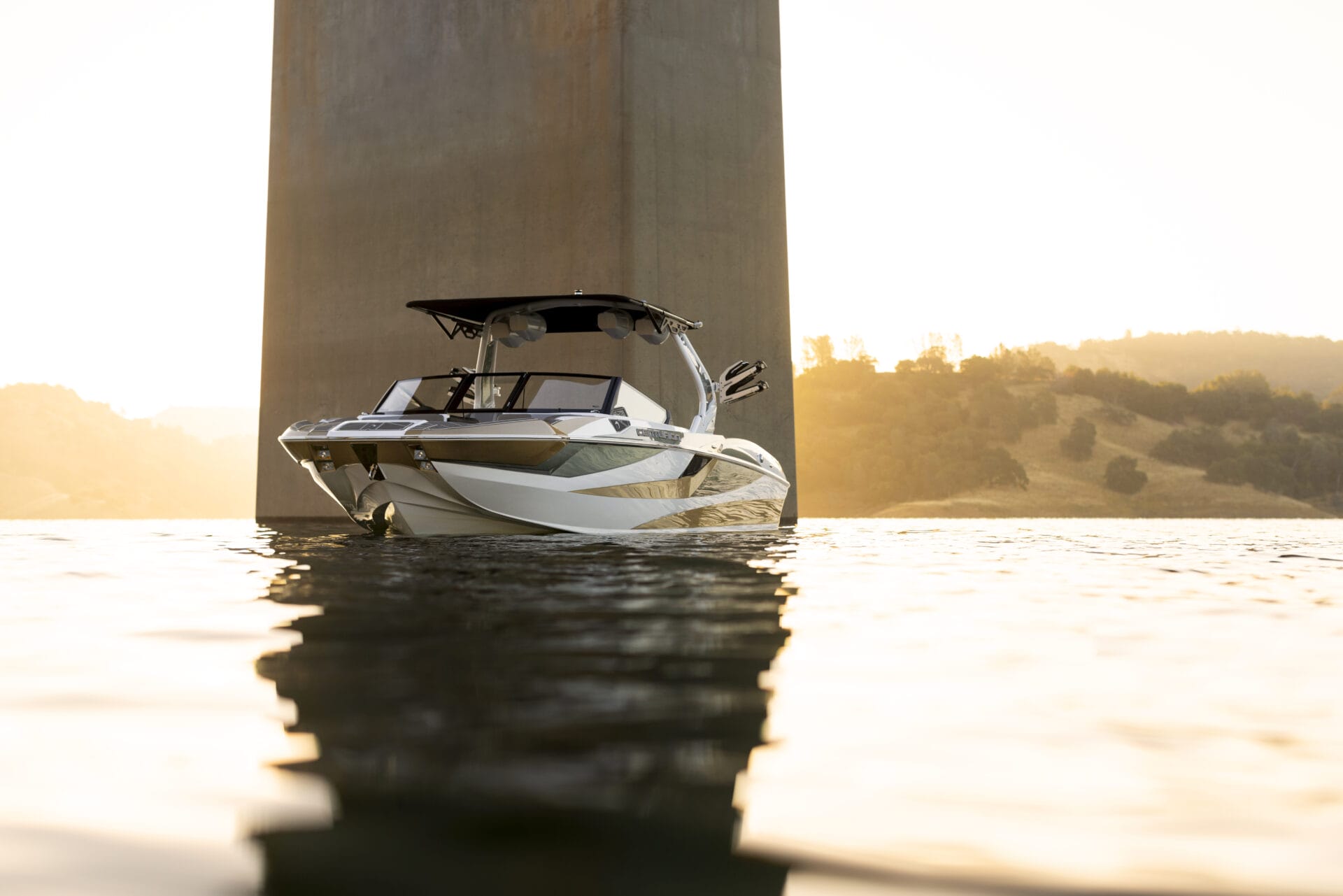 A motorboat anchored underneath a tall bridge pillar, with calm water reflecting the sunset and distant hills in the background.