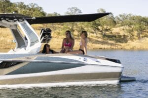 Three women sit and chat on a boat in a lake with dry, grassy hills and trees in the background.