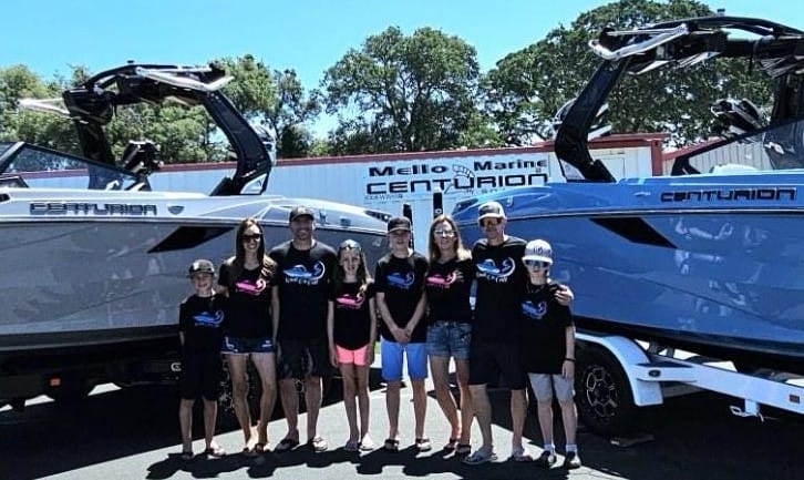 A group of nine people, including adults and children, stand in front of two parked Centurion boats outside a Mello Marine dealership. They are all wearing matching T-shirts.