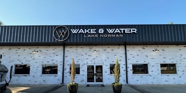 Storefront of Wake & Water Lake Norman with a black and white exterior brick wall, double glass doors, two planter pots with trees on either side, and a clear blue sky.
