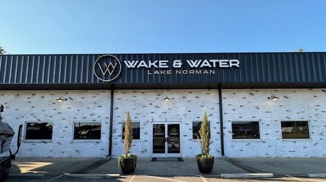 Storefront of Wake & Water Lake Norman with a black and white exterior brick wall, double glass doors, two planter pots with trees on either side, and a clear blue sky.