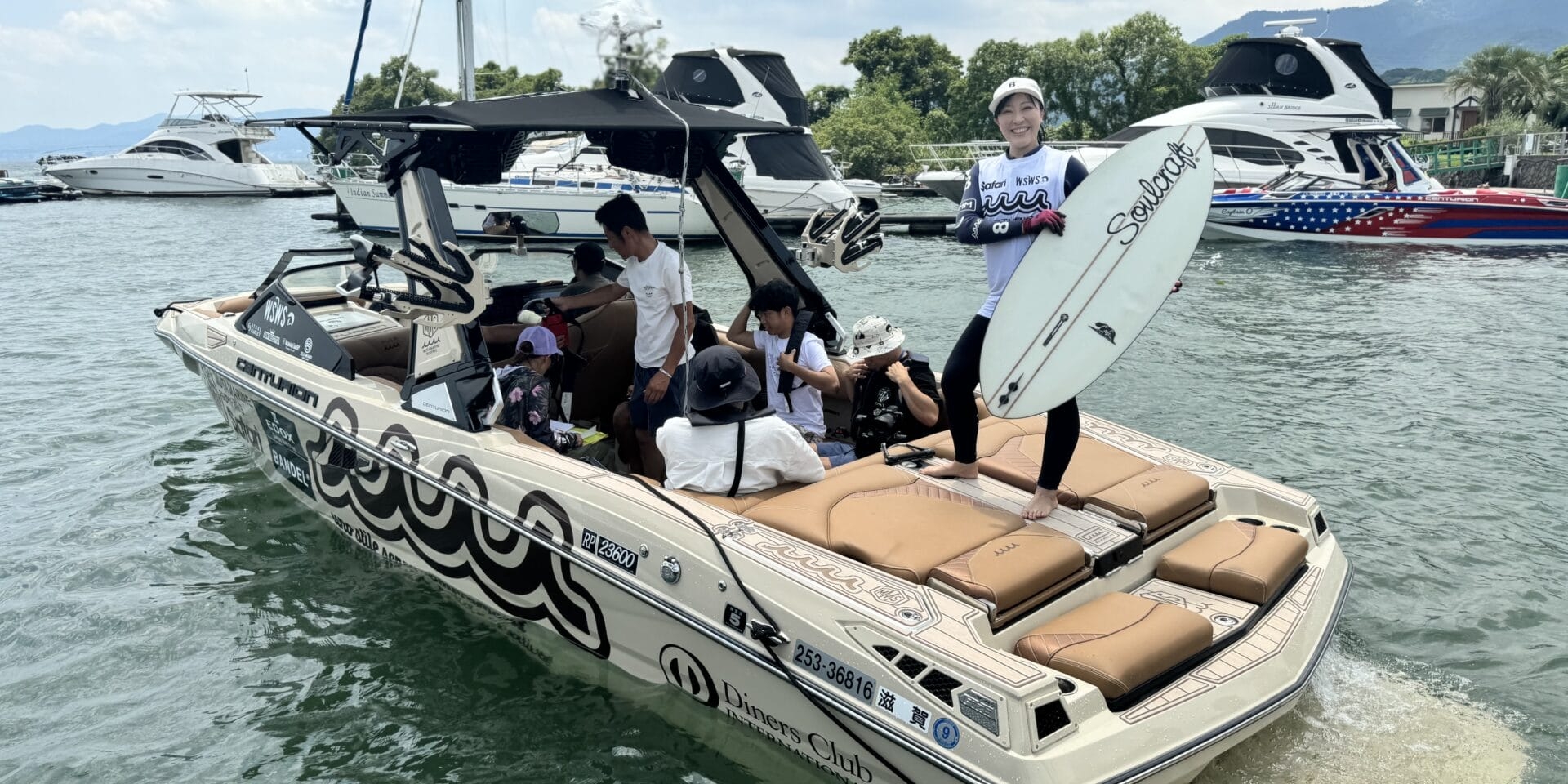 Group of people on a motorboat at a marina, one person standing with a surfboard. Other boats are visible in the background.