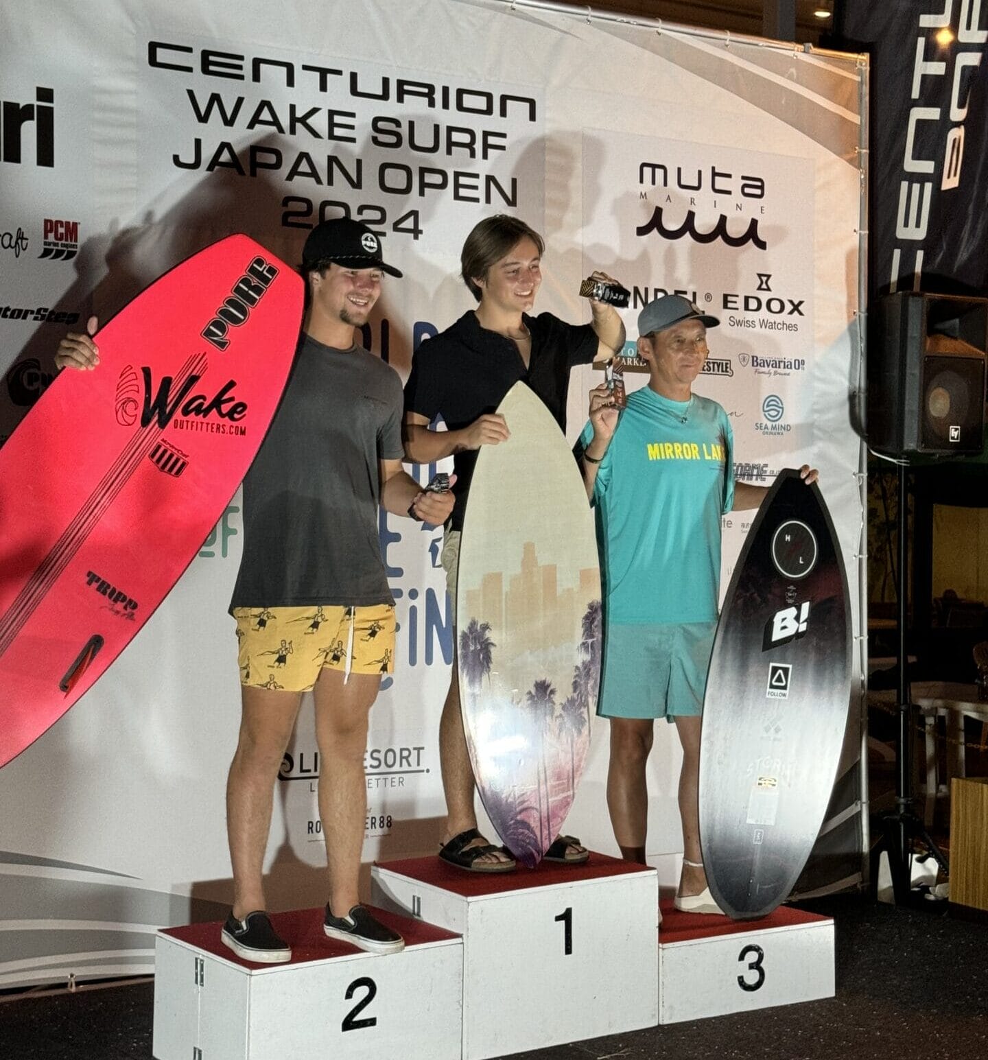 Three people stand on a winners' podium at the Centurion Wake Surf Japan Open 2024, each holding surfboards and trophies. The backdrop features event and sponsor logos.