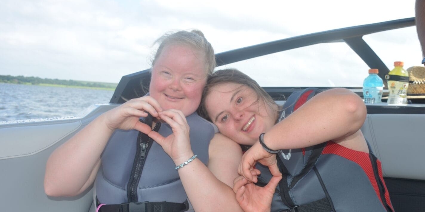 Two people wearing life jackets sit on a boat, making heart shapes with their hands. A third person stands nearby, and drinks are visible in the background.