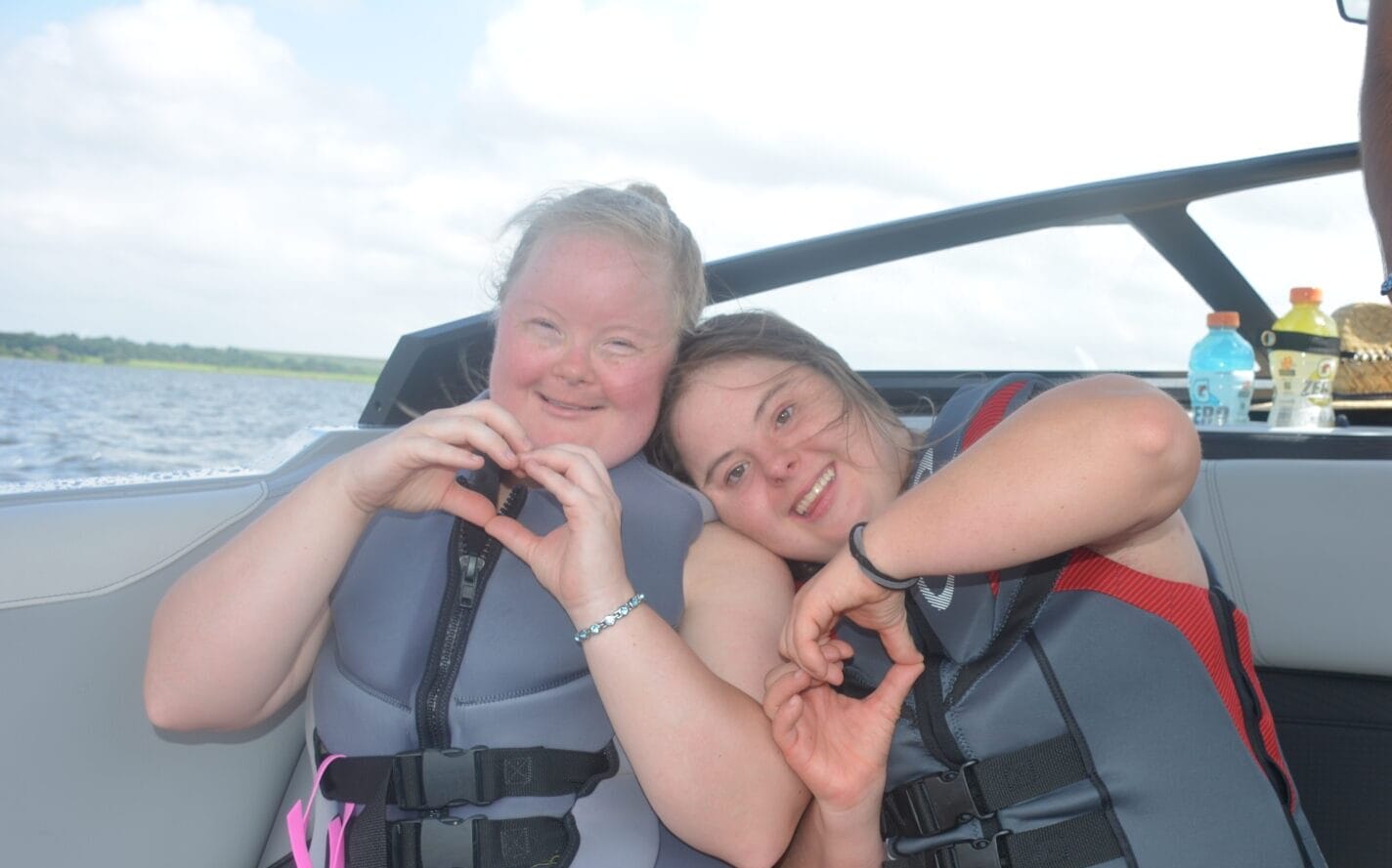 Two people wearing life jackets sit on a boat, making heart shapes with their hands. A third person stands nearby, and drinks are visible in the background.