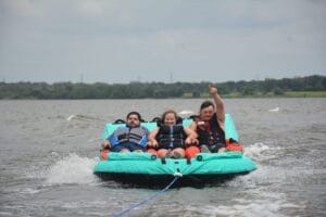 Three people wearing life jackets are seated on an inflatable towable tube on the water, smiling and raising a hand in the air. Trees and a cloudy sky are visible in the background.
