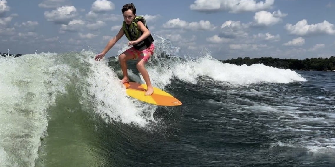 A person wearing a life vest rides a yellow surfboard on a wave under a blue sky with scattered clouds.