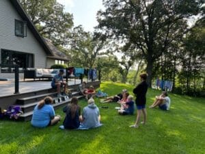 A group of people sit on and around the steps of a house's deck, engaged in conversation. Towels and clothes hang dry on the railing of the deck. The scene is set in a grassy area with trees.