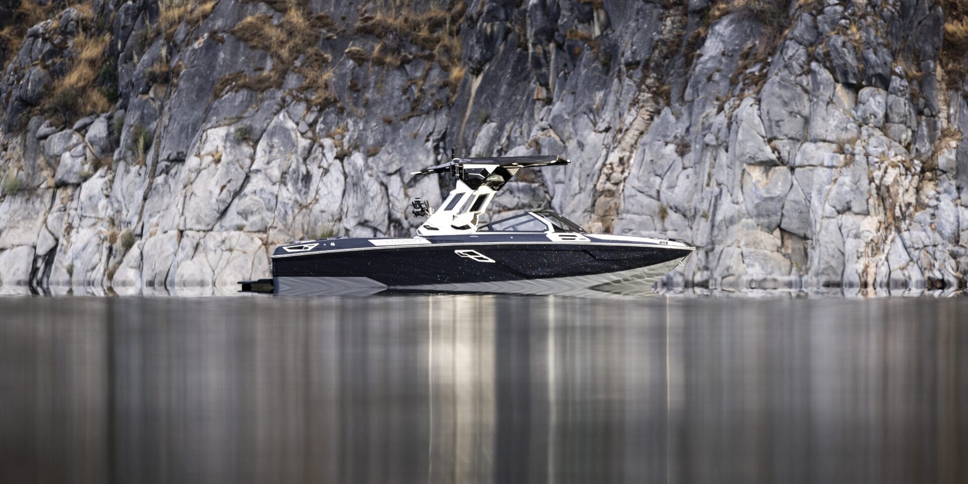 A black and white speedboat is anchored near a rocky cliff shoreline, partially reflected in the calm water.