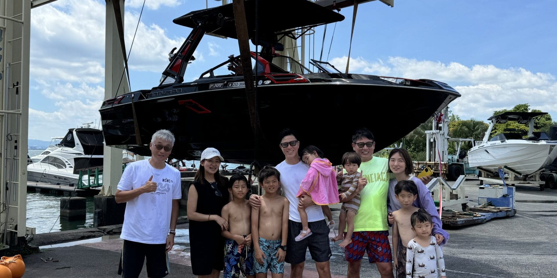 A group of people, including men, women, and children, stand together in front of a large boat suspended in the air by a mechanical lift at a marina.