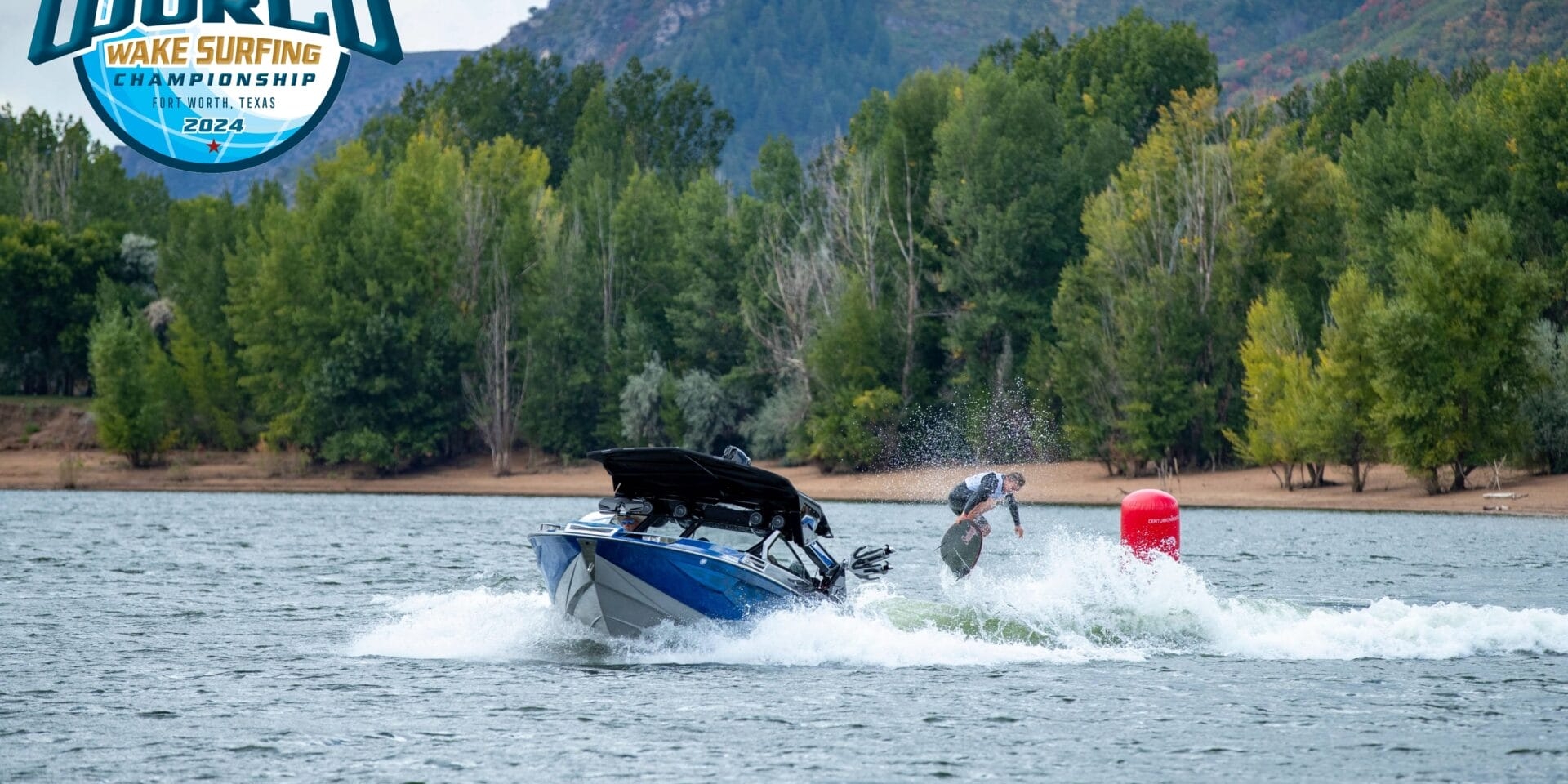A person performs a wake surfing trick behind a speedboat on a lake with a forested hillside in the background. A logo indicates 