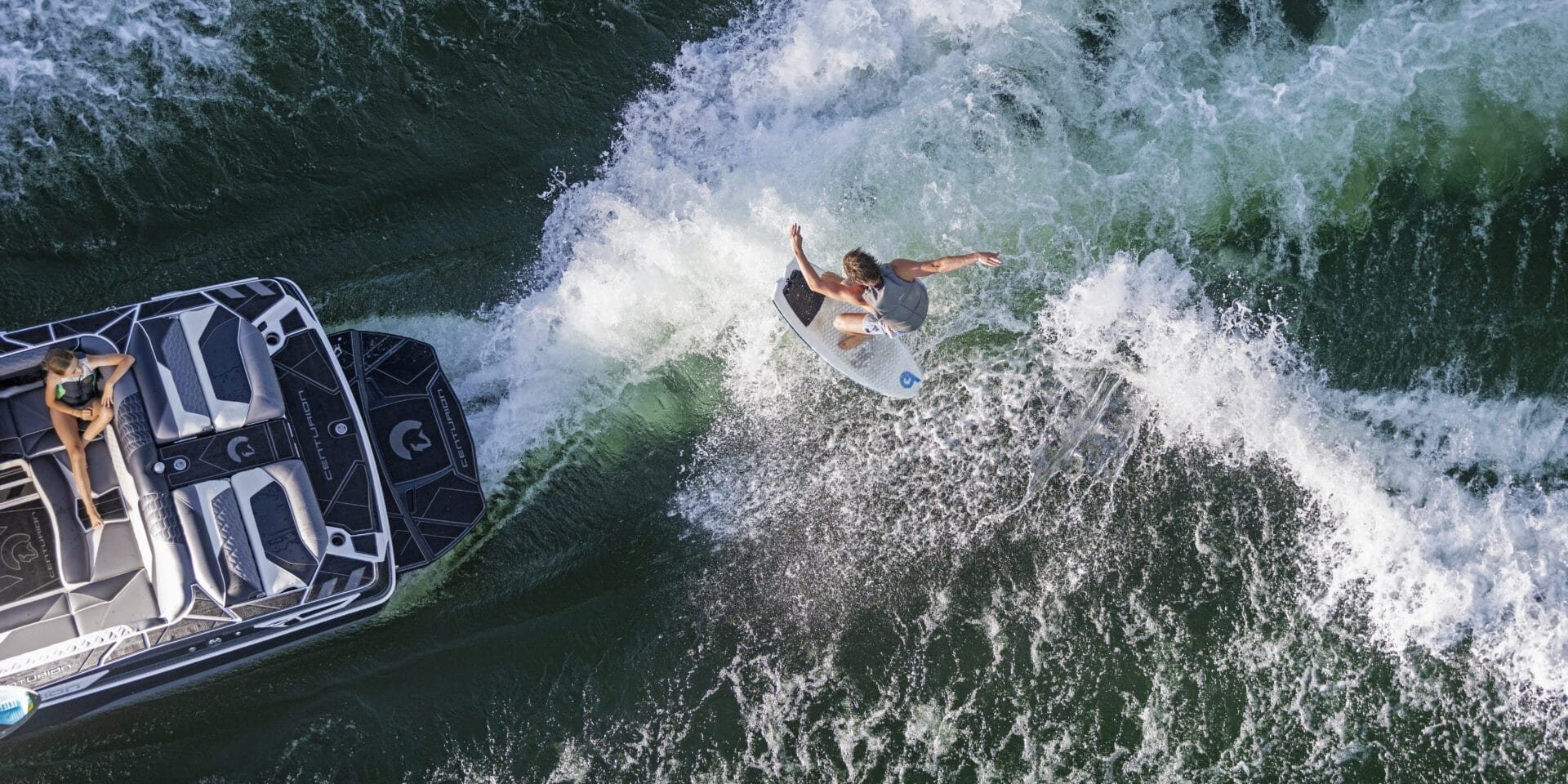 A person surfs the wake behind a speedboat on a green body of water.
