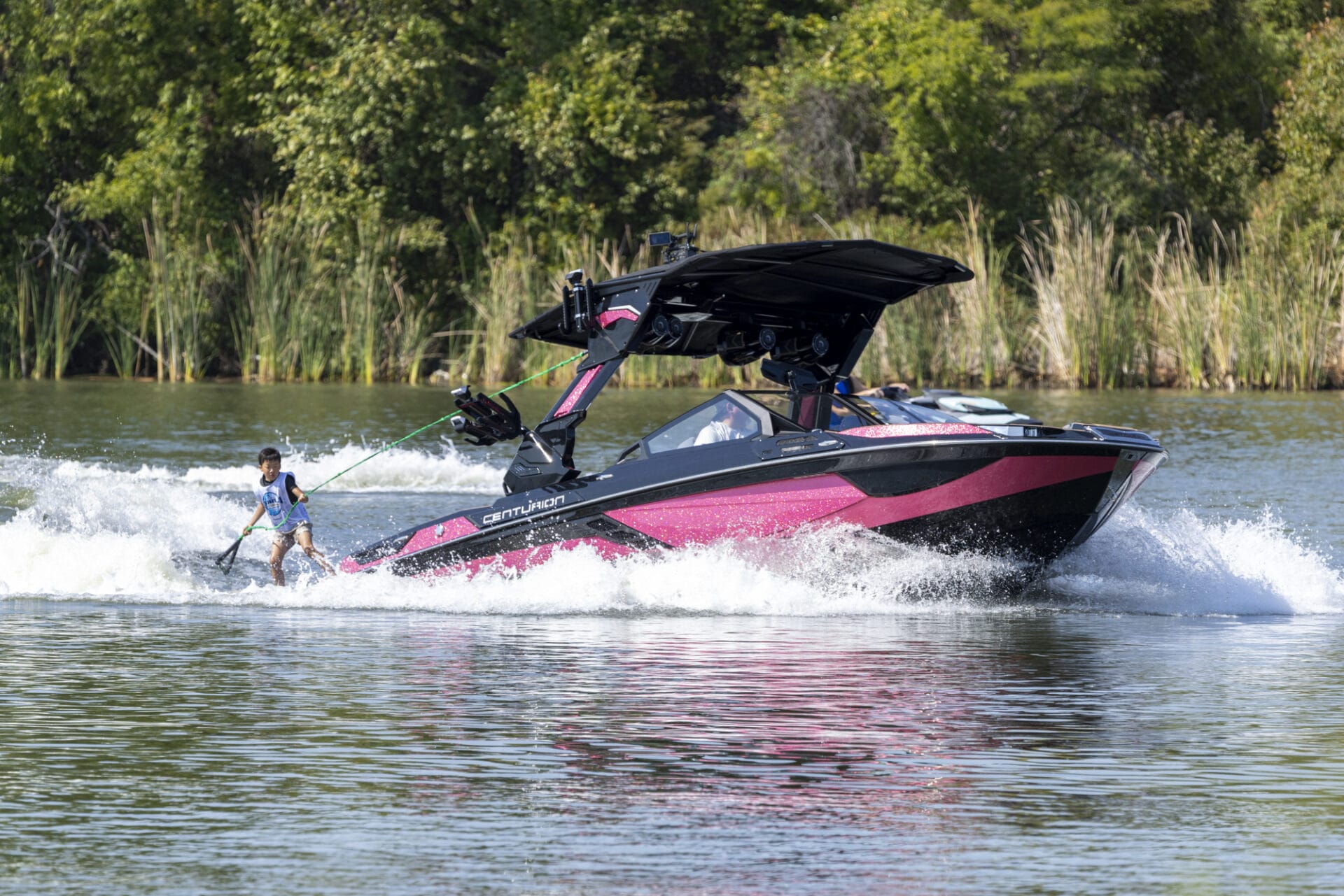 A person is wakeboarding on a lake, being towed by a pink and black boat. Trees and reeds line the background.