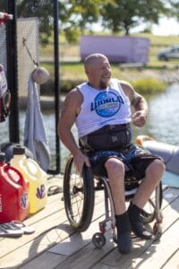 A man in a wheelchair smiles near a body of water. He's wearing a tank top and shorts, and there are gas cans nearby.
