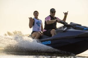 Two people riding a Sea-Doo jet ski on the water, wearing life jackets, with both making a hand gesture.