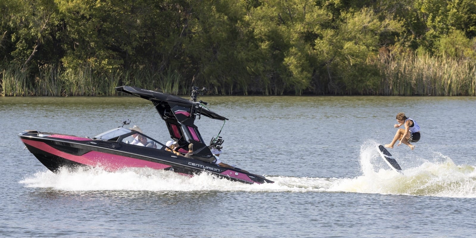 A person is wakeboarding behind a speedboat on a lake with trees in the background.