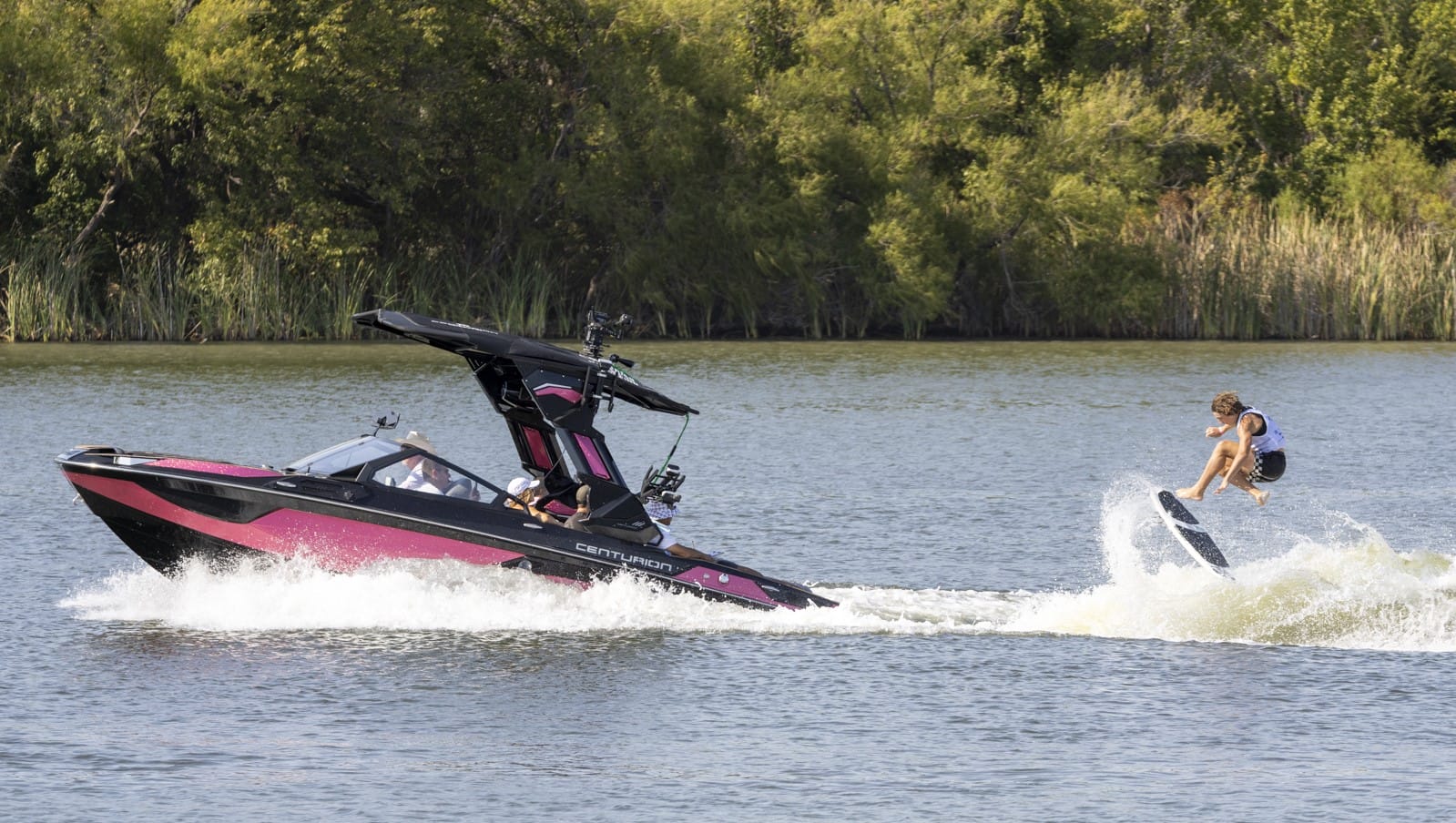 A person is wakeboarding behind a speedboat on a lake with trees in the background.
