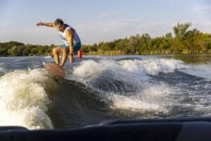 Person surfing on a river wave, wearing a blue tank top and shorts. Trees and a clear sky are in the background.