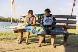 Two individuals sit on a bench beside a body of water, each holding a wakeboard, with gear and flags nearby.