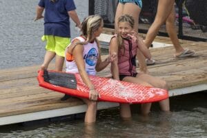 Two people sitting on a dock, holding a red surfboard, with others walking nearby. Water is visible in the background.