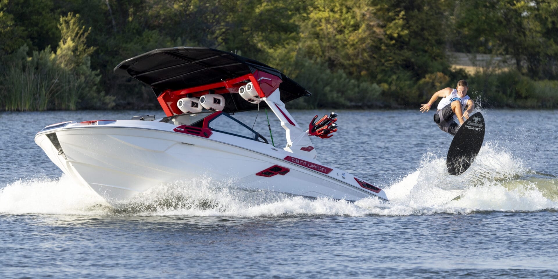 A person wakeboarding performs a trick in the air beside a moving motorboat on a lake with trees in the background.