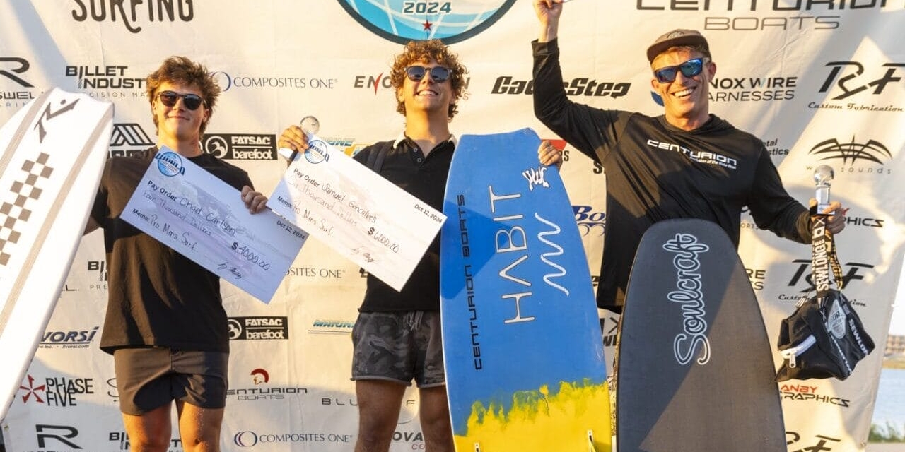 Three people stand on a podium holding surfboards and prize checks at a wake surfing championship event.