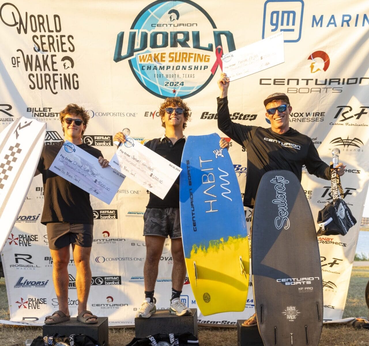 Three people stand on a podium holding surfboards and prize checks at a wake surfing championship event.