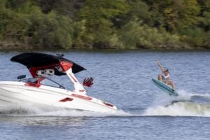 A person is wakeboarding behind a red and white boat on a lake, performing a jump with one arm raised. Trees are visible in the background.