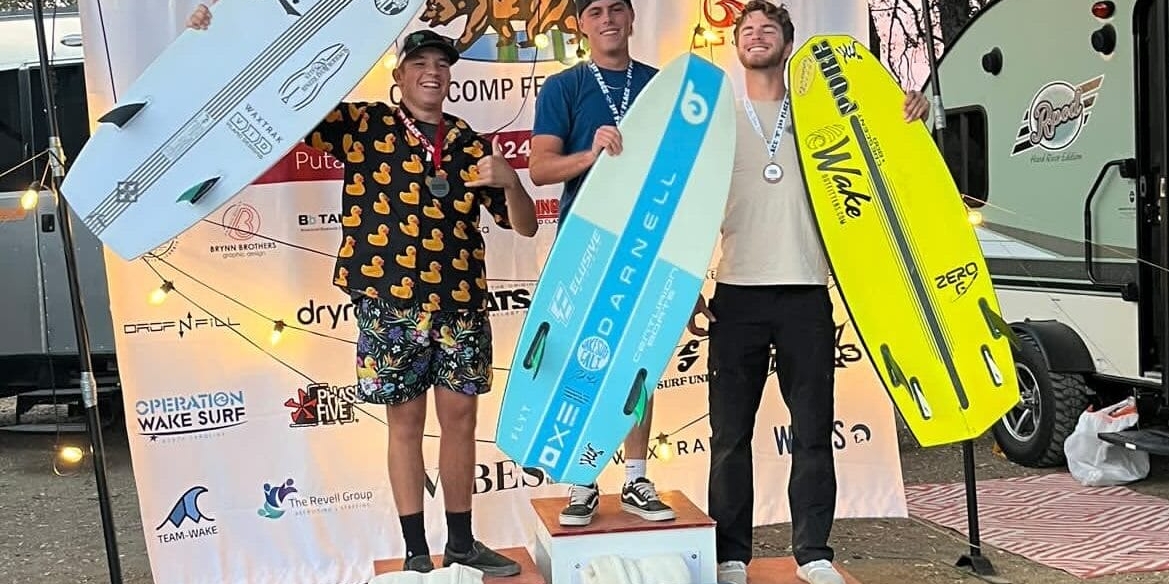 Three people stand on podiums holding large wakeboards in front of a branded backdrop at a competition event.