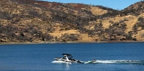 A person water skiing on a lake behind a boat on a sunny day, with dry, rocky hills in the background.
