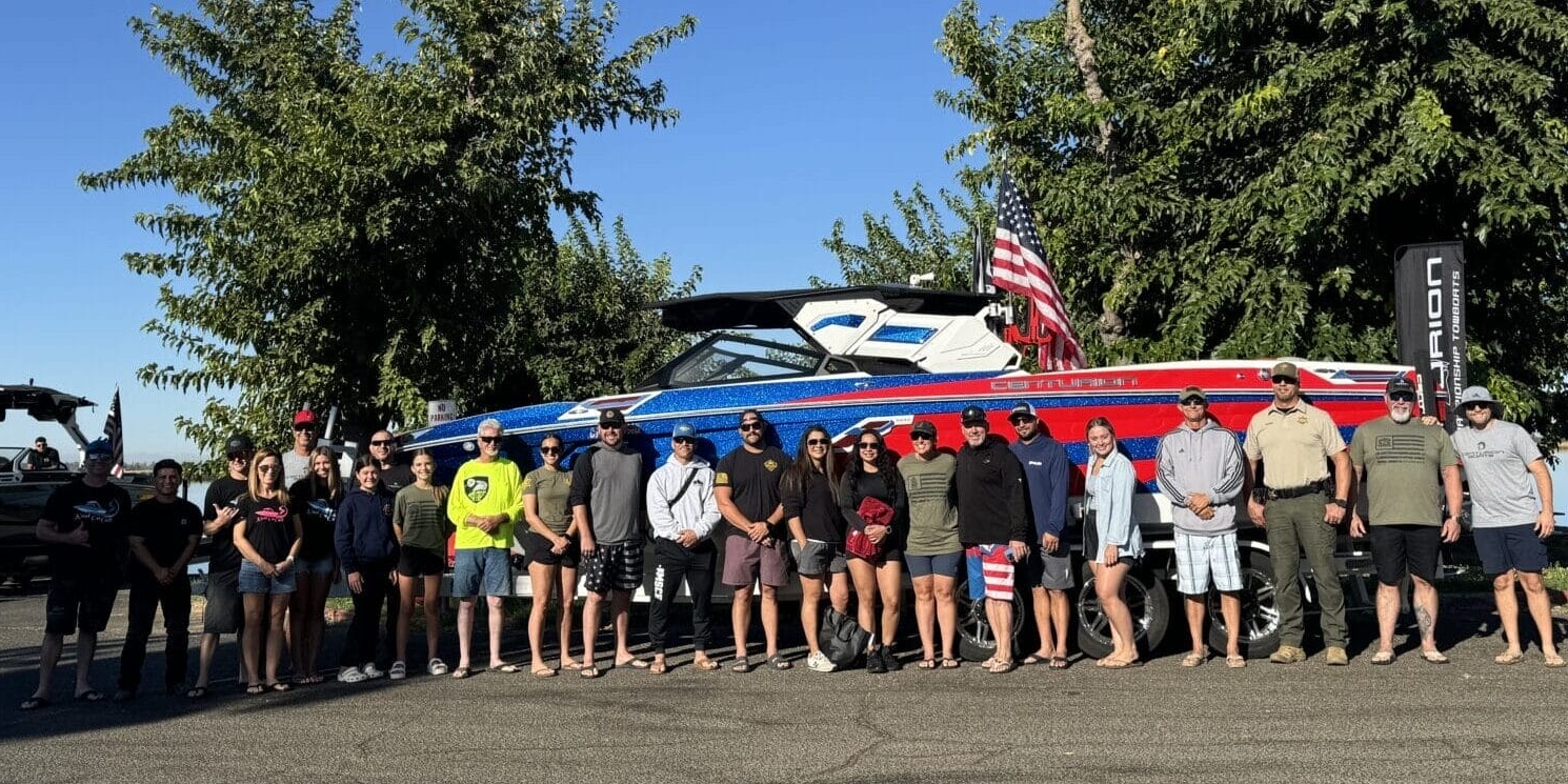 A group of people stands in front of a large blue and red boat on a trailer, with trees in the background and a clear blue sky above.